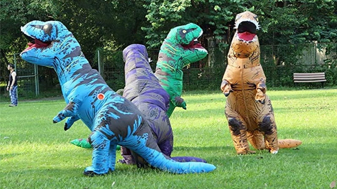 People in colorful inflatable dinosaur costumes running on a grassy park field. Fun outdoor event.